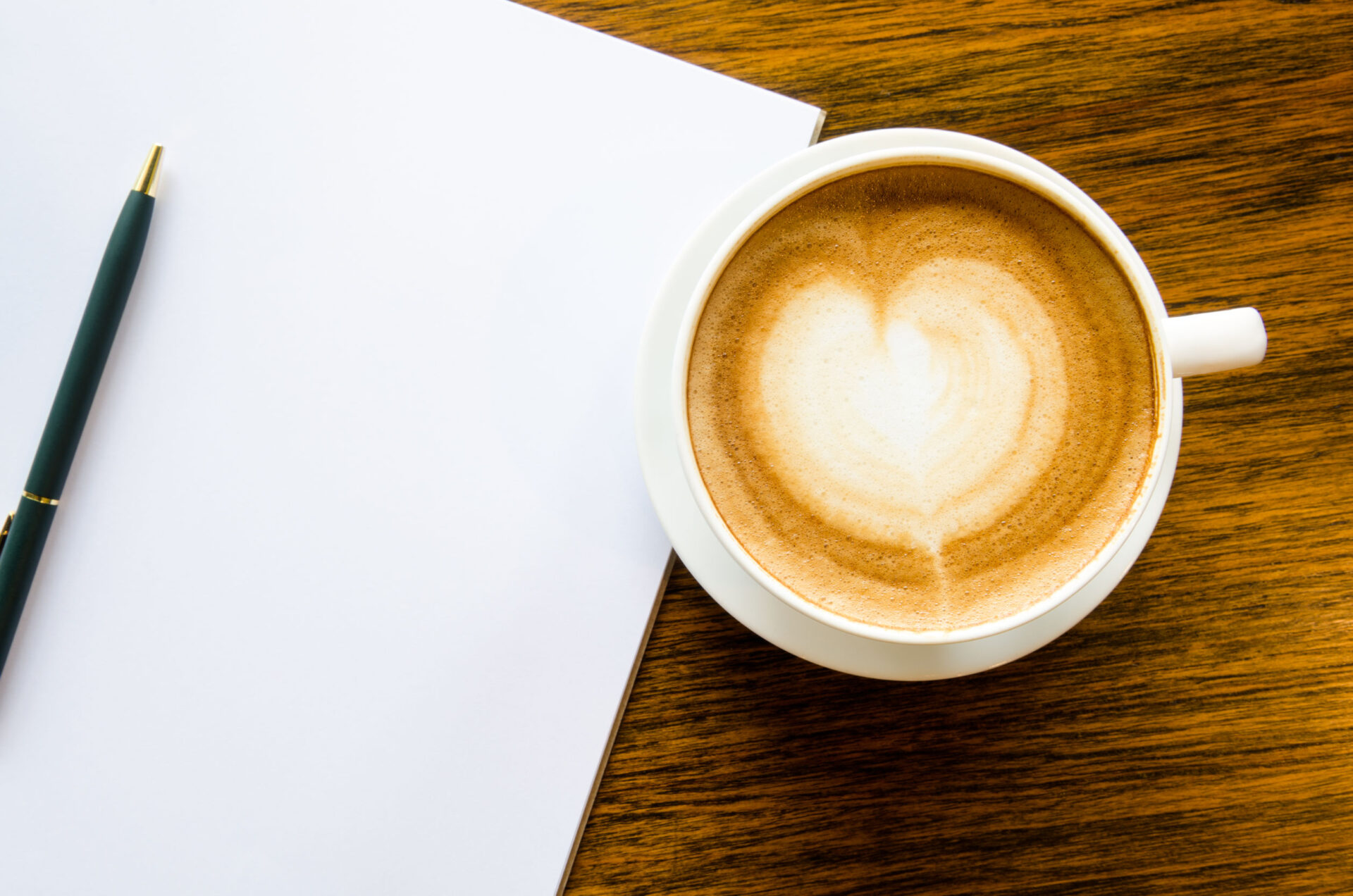 A cup of coffee with heart shape, pen and open blank book on wood background